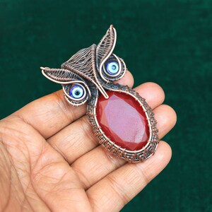 Może przedstawiać: Wisiorek w kształcie sowy z drutu miedzianego z czerwonym kamieniem szlachetnym i niebieskimi detalami złego oka.