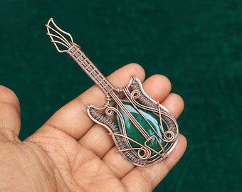 May include: A copper wire guitar pendant featuring a green gemstone at its centre. The pendant is intricately crafted with wirework and is held in a hand.