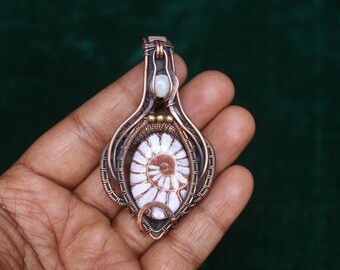 May include: A handcrafted pendant featuring a fossilized ammonite shell encased in copper wire. The pendant has a spiral design in shades of white and brown, with a copper wire frame and a small, white stone at the top.