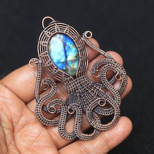 Könnte beinhalten: Ein Anhänger aus Kupferdraht, der eine Octopus-Form hat und einen blauen und grünen Labradorit-Edelstein in der Mitte trägt.