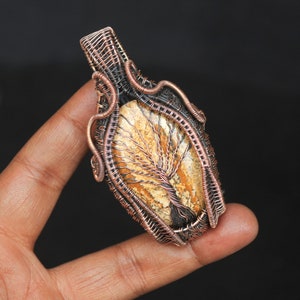 May include: A copper wire wrapped pendant featuring a tree of life design in a brown and beige stone.