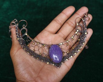 May include: A handcrafted copper and brass necklace featuring a large, oval, polished purple gemstone. The intricate metalwork includes delicate wire mesh and small gold-colored accents, creating a unique and artistic design.