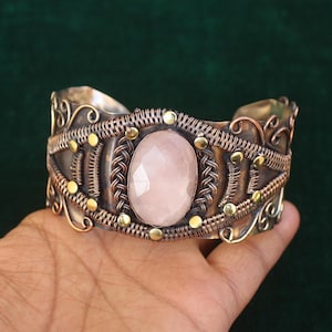May include: A handcrafted cuff bracelet with a large, oval rose quartz stone at the center. The silver-toned metal is intricately detailed with copper wire and small gold-colored accents. The bracelet is a wide band, designed to be worn on the wrist.