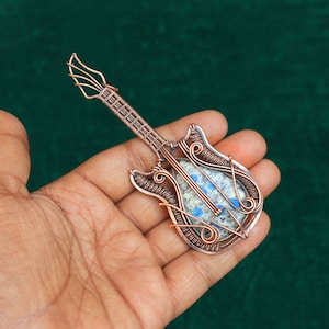 May include: A copper wire guitar pendant with a blue and white stone inlay. The pendant is held in a hand.