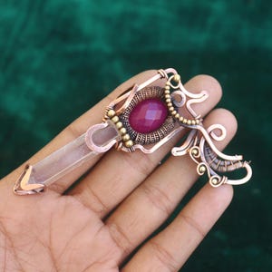 May include: A handcrafted copper pendant featuring a clear quartz crystal and a large, oval, deep pink gemstone. The pendant has intricate wirework and small gold-colored beads. The design is held in a hand against a dark green background.