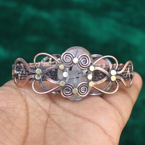 May include: A copper wire wrapped bracelet featuring a clear quartz stone at its centre. The bracelet is adorned with small gold beads and wire swirls.