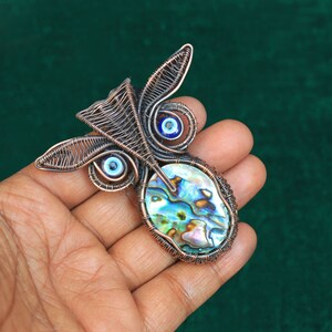 Może przedstawiać: Wisiorek w kształcie sowy z drutu miedzianego z muszlą abalone i niebieskimi oczami.