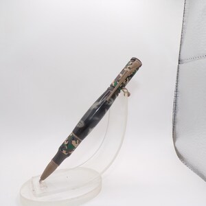 May include: A camouflage-patterned pen with a silver clip and a brown tip. The pen is displayed on a clear acrylic stand against a white background. The pen's design incorporates shades of green, brown, and black.