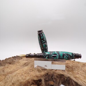 Pu&ograve; includere: Una penna stilografica con un design marmorizzato nero, verde acqua e grigio. La penna &egrave; aperta, rivelando un pennino dorato. La penna poggia su un supporto in acrilico trasparente, su uno sfondo di legno naturale.