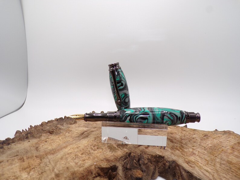 Pu&ograve; includere: Una penna stilografica con un design marmorizzato nero, verde acqua e grigio. La penna &egrave; aperta, rivelando un pennino dorato. La penna poggia su un supporto in acrilico trasparente, su uno sfondo di legno naturale.