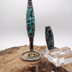 Pu&ograve; includere: Una penna artigianale con un design marmorizzato verde acqua, nero e bianco. La penna &egrave; esposta su un supporto argentato con un portapenne in acrilico trasparente. Il cappuccio della penna &egrave; posto accanto alla penna. La penna ha una finitura in metallo scuro.