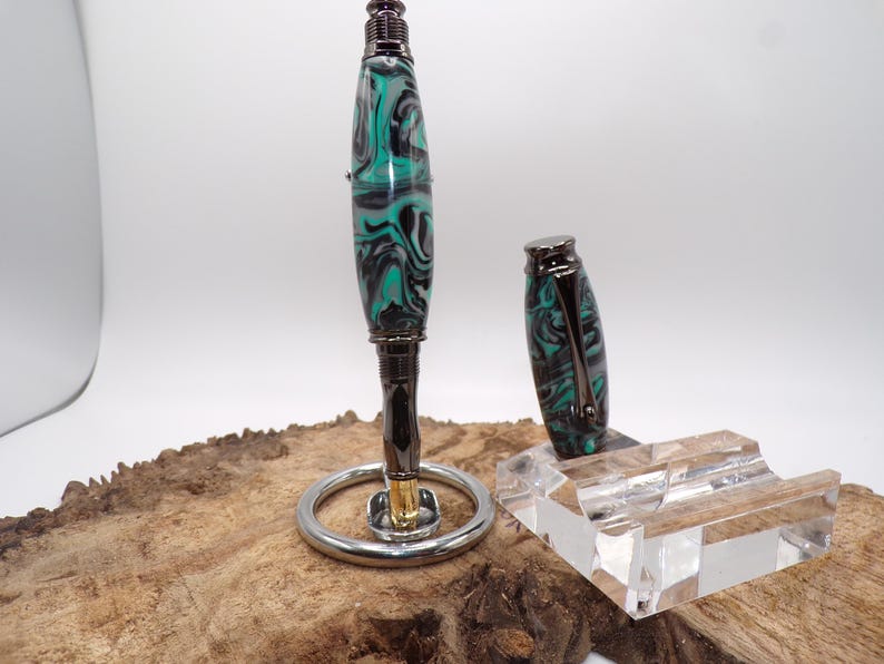 Pu&ograve; includere: Una penna artigianale con un design marmorizzato verde acqua, nero e bianco. La penna &egrave; esposta su un supporto argentato con un portapenne in acrilico trasparente. Il cappuccio della penna &egrave; posto accanto alla penna. La penna ha una finitura in metallo scuro.