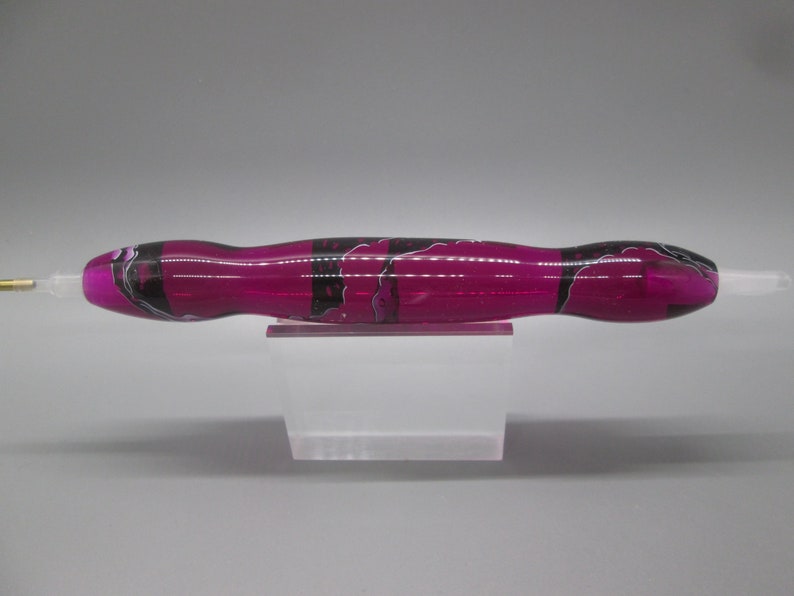 May include: A pink and black marbled pen with a clear tip. The pen is sitting on a clear acrylic stand.