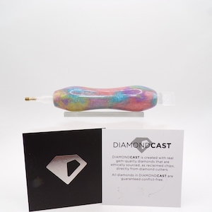 May include: A close-up of a colorful diamond drill pen with a clear plastic tip. The pen is made with real, ethically sourced diamonds. The text on the card reads "DIAMONDCAST is created with real gem-quality diamonds that are ethically sourced, as reclaimed chips, directly from diamond cutters. All diamonds in DIAMONDCAST are guaranteed conflict-free."