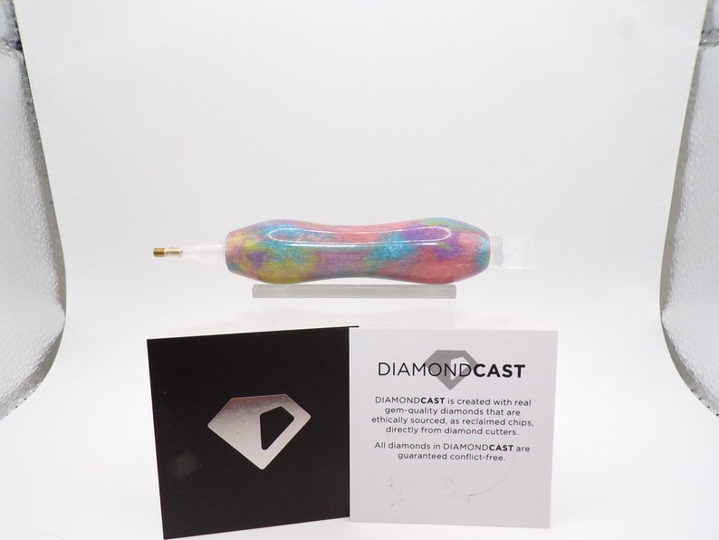 May include: A close-up of a colorful diamond drill pen with a clear plastic tip. The pen is made with real, ethically sourced diamonds. The text on the card reads "DIAMONDCAST is created with real gem-quality diamonds that are ethically sourced, as reclaimed chips, directly from diamond cutters. All diamonds in DIAMONDCAST are guaranteed conflict-free."