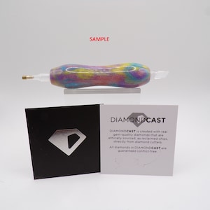 May include: A close-up of a colorful, swirl-patterned, diamond-encrusted pen with a gold tip. The pen is on a white surface with a black and white card that reads "DIAMONDCAST is created with real gem-quality diamonds that are ethically sourced, as reclaimed chips, directly from diamond cutters. All diamonds in DIAMONDCAST are guaranteed conflict-free."