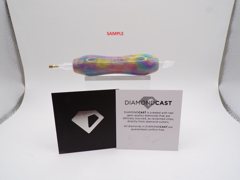 May include: A close-up of a colorful, swirl-patterned, diamond-encrusted pen with a gold tip. The pen is on a white surface with a black and white card that reads "DIAMONDCAST is created with real gem-quality diamonds that are ethically sourced, as reclaimed chips, directly from diamond cutters. All diamonds in DIAMONDCAST are guaranteed conflict-free."