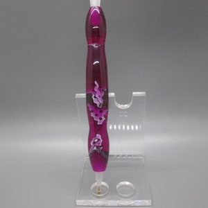 May include: A pink and white patterned pen with a clear top and a gold tip, displayed on a clear acrylic stand.