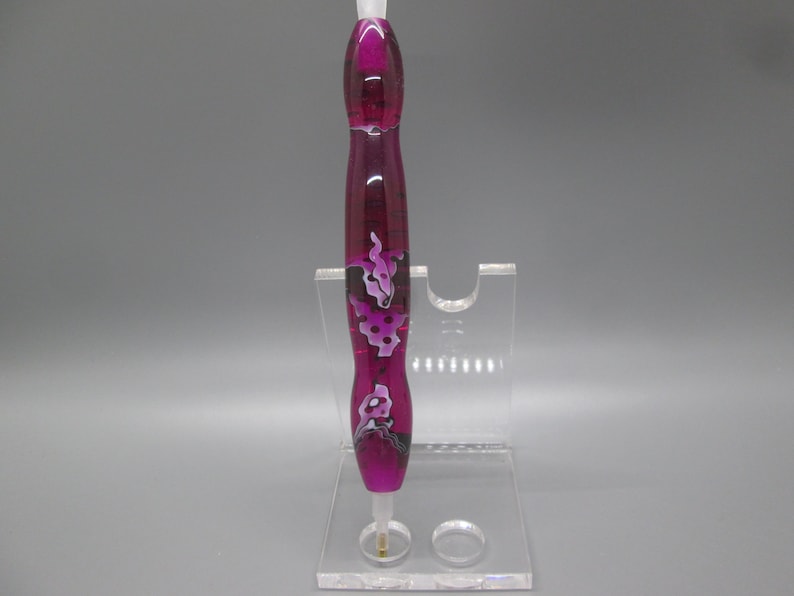 May include: A pink and white patterned pen with a clear top and a gold tip, displayed on a clear acrylic stand.