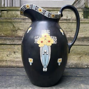 May include: Black ceramic pitcher with a floral design featuring yellow flowers and blue and green accents. The pitcher has a wide mouth and a handle.