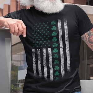 May include: A black t-shirt with a distressed American flag design featuring green shamrocks. The flag is white and grey with a green shamrock design.