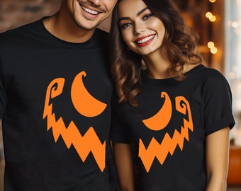 Puede incluir: Una pareja lleva camisetas negras a juego con un diseño de cara de calabaza naranja. El diseño es un simple contorno de una cara de calabaza con una amplia sonrisa y dos ojos.