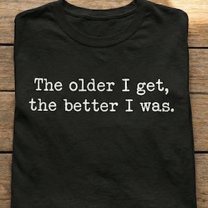 Peut inclure: T-shirt noir avec le texte blanc "The older I get, the better I was." Le t-shirt est fait d'une matière douce et est posé à plat sur une surface en bois. Le texte est centré sur le t-shirt.