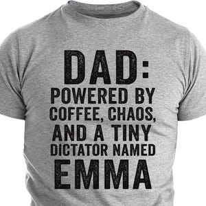 以下が含まれることがあります： 「DAD: POWERED BY COFFEE, CHAOS, AND A TINY DICTATOR NAMED EMMA」と黒い太字で書かれた、グレーのTシャツ。Tシャツは半袖で、クルーネックです。