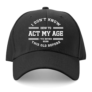 Puede incluir: Gorra de béisbol negra con el texto blanco: "I don't know how to act my age, I've never been this old before". La gorra tiene una visera curva y un botón en la parte superior.