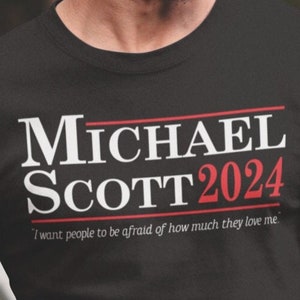 May include: Black t-shirt with white text that reads "MICHAEL SCOTT 2024" and the smaller text "I want people to be afraid of how much they love me."