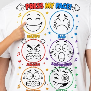 Pode incluir: Camiseta branca com gráficos coloridos de rostos emoji representando diferentes emoções. O texto "PRESS MY FACE!" está no topo e "YOU'RE IN CONTROL" na parte inferior. Uma criança aponta para a camiseta.