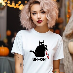 Könnte beinhalten: Weißes T-Shirt mit einer schwarzen Katze, die ein blutiges Messer hält. Der Text "UH-OH!" ist unter der Katze aufgedruckt.