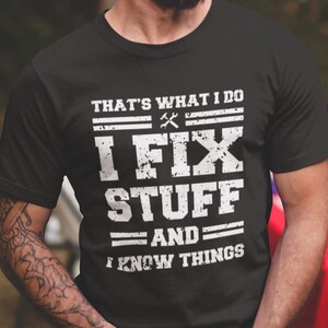 May include: Black t-shirt with white text that reads "THAT'S WHAT I DO, I FIX STUFF, AND I KNOW THINGS." The text includes a wrench and hammer graphic. The shirt is short-sleeved.