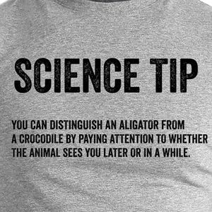 May include: A grey t-shirt featuring the words "SCIENCE TIP" in bold black font. Underneath, a humorous science fact explains how to tell alligators and crocodiles apart. The text is also in black.