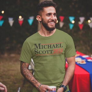 May include: A green t-shirt with the text "MICHAEL SCOTT 2024" in black and red. The text "I  expect people to be afraid of how much they love me" is printed in black below the main text.