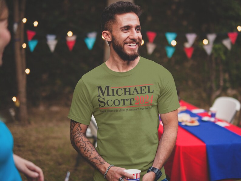 May include: A green t-shirt with the text "MICHAEL SCOTT 2024" in black and red. The text "I  expect people to be afraid of how much they love me" is printed in black below the main text.