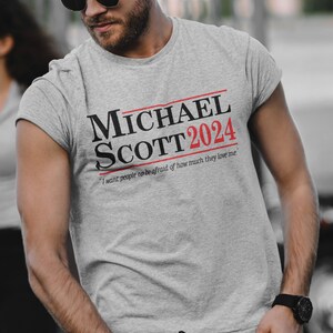 May include: A grey t-shirt with a black and red print that says "MICHAEL SCOTT 2024" and "I want people to be afraid of how much they love me."