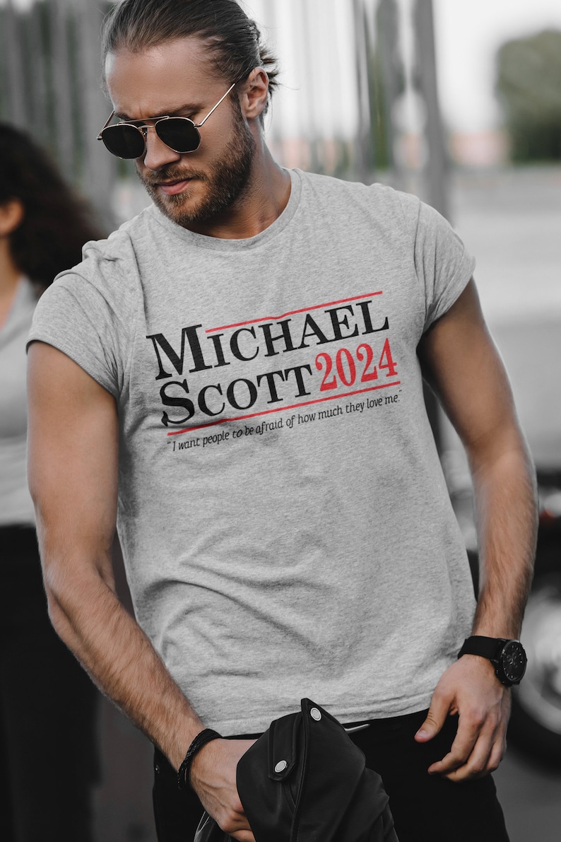 May include: A grey t-shirt with a black and red print that says "MICHAEL SCOTT 2024" and "I want people to be afraid of how much they love me."