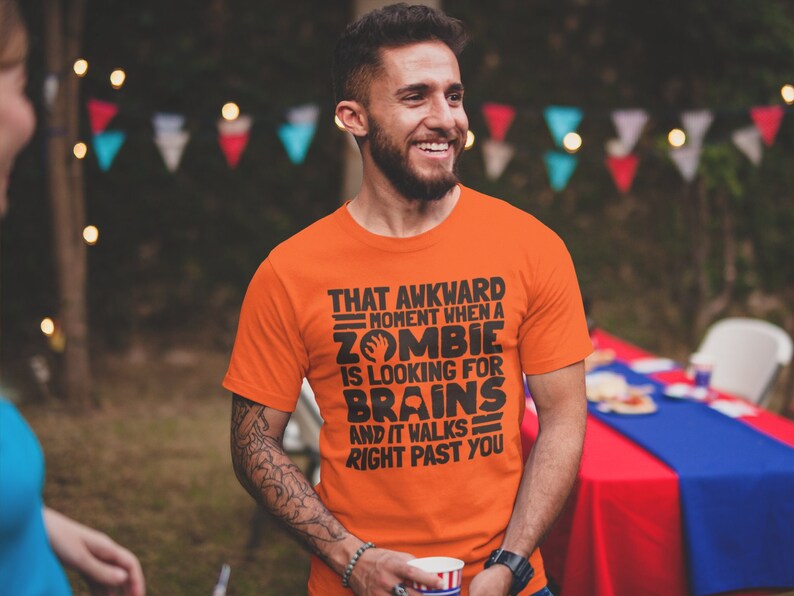 May include: A man wearing an orange t-shirt with a humorous graphic that reads "That awkward moment when a zombie is looking for brains and it walks right past you".