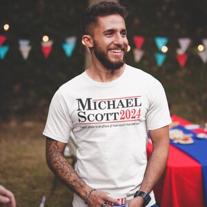 May include: White t-shirt with the text "MICHAEL SCOTT 2024" in black and red. Below the text is the phrase "I want people to be afraid of how much they love me."
