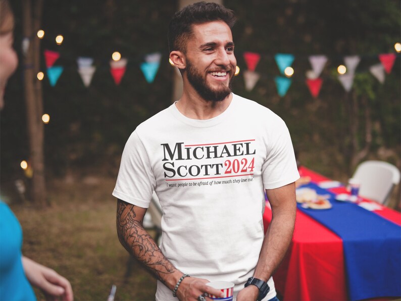 May include: White t-shirt with the text "MICHAEL SCOTT 2024" in black and red. Below the text is the phrase "I want people to be afraid of how much they love me."