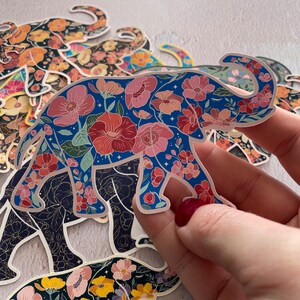 K&ouml;nnte beinhalten: Ein blauer Elefanten-Aufkleber mit einem Blumenmuster. Der Elefant ist umgeben von anderen Blumenaufklebern, darunter ein schwarzer Elefant mit einem Blumenmuster.