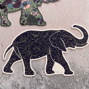 K&ouml;nnte beinhalten: Schwarzer und goldener Elefanten-Silhouetten-Aufkleber mit floralem Muster. Der Elefant geht nach rechts und hat seinen R&uuml;ssel erhoben. Das Blumenmuster ist filigran und detailliert, mit kleinen Blumen und Bl&auml;ttern.