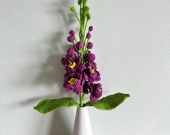 May include: A handcrafted crochet flower stalk featuring vibrant purple petals with small yellow centers, attached to a bright green stem with textured leaves, displayed in a smooth, white, conical-shaped ceramic vase.