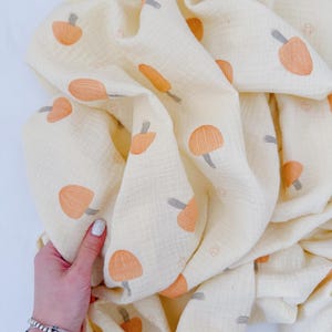 Könnte beinhalten: Cremefarbene Babydecke mit einem Muster aus orangefarbenen Pilzen und grauen Stielen. Der weiche, leichte Stoff ist gefaltet und zeigt das Pilzdesign. Die Decke wird von einer Hand mit silbernen Armbändern gehalten.