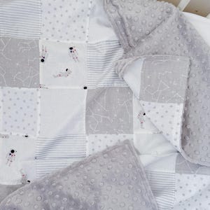 May include: A patchwork baby blanket with a space theme. The blanket features squares of white, grey, and patterned fabric. Some squares have astronauts, constellations, and polka dots. The border is a soft, grey, dimpled fabric.