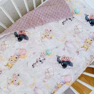 May include: A baby blanket with a whimsical print of animals, hot air balloons, and celestial elements in soft pastel colors. The blanket has a textured, mauve-colored backing. The blanket is placed inside a white crib.