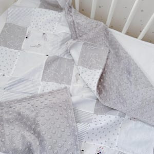 Op de afbeelding: Een patchwork babydeken in een wieg. De deken heeft vierkanten van witte, grijze en bedrukte stoffen, waaronder strepen, stippen en sterrenbeeldontwerpen. De achterkant en rand zijn van een zachte, grijze stof met nopjes.