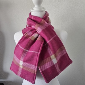 May include: A vibrant pink plaid scarf displayed on a mannequin. The scarf features a classic plaid pattern in shades of pink, white, and tan. The soft, warm material suggests a cozy accessory for colder weather.
