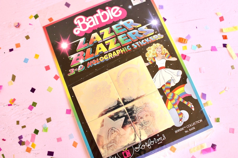 May include: A package of Barbie Lazer Blazers 3-D Holographic Stickers. The package is black with a rainbow design and features a holographic image of Barbie wearing a white dress and rainbow striped tights. The package includes four holographic stickers.